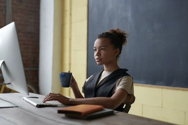 View of young woman seated at desk learning on desktop in front of chalkboard and holding a mug