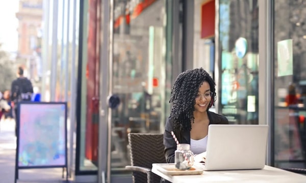 Smiling woman seated at outdoor table beside store window, working on laptop with drink beside her