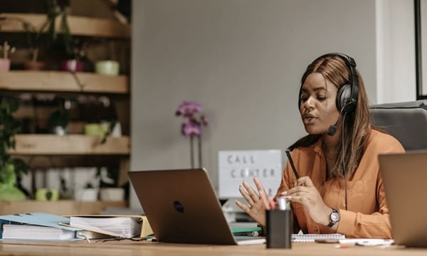 View of employee seated at desk in front of laptop wearing headphones in the middle of a call, gesticulating and speaking