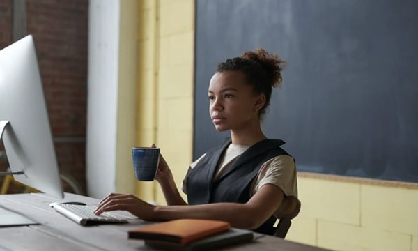 View of young woman seated at desk learning on desktop in front of chalkboard and holding a mug