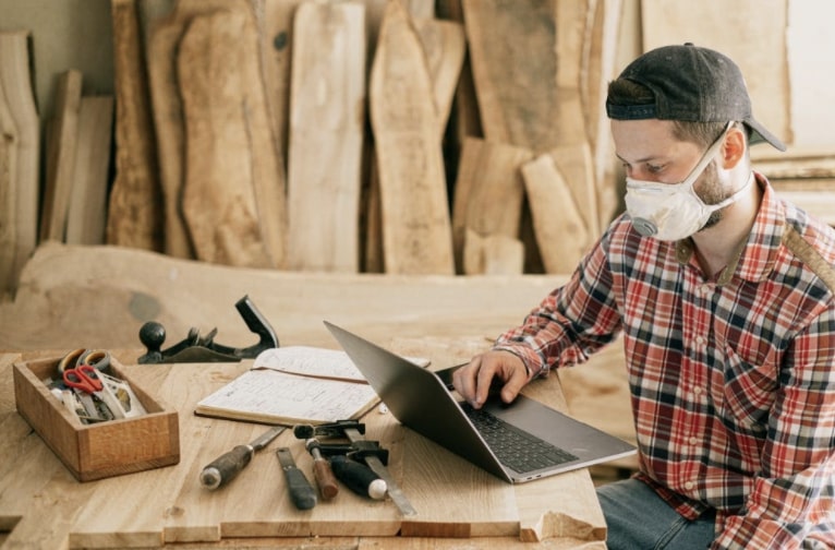 A carpenter working on laptop in a shop surrounded by woodworking toolbox and tools in foreground and wood in background