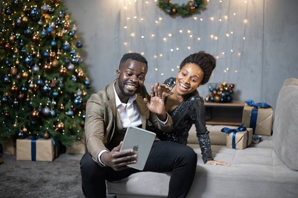 Happy couple celebrating the holidays at home with decorations in the background, and using a tablet to video chat with others