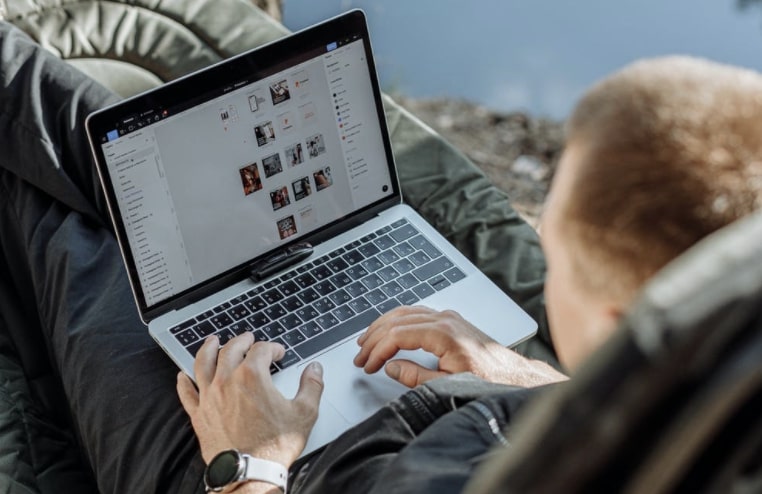 Over the shoulder view of casual-looking man working on laptop in a reclined position on cushion outdoors