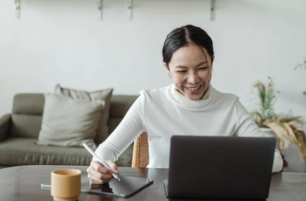 Smiling happy woman seated at desk interacting on digital pad while talking on laptop at home