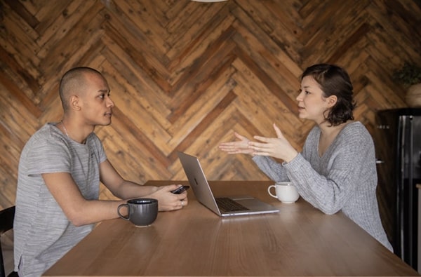 View of 1-1 conversation between woman talking with open laptop and gesticulating, and man listening intently in communal work space