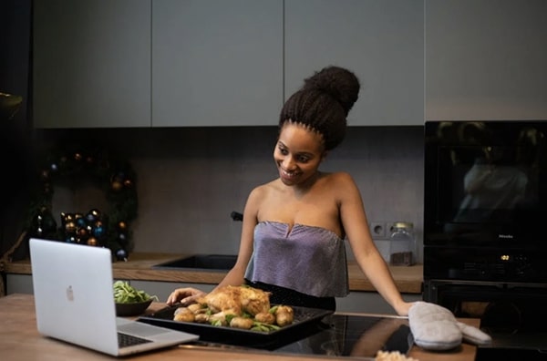 Woman at home in stylish kitchen video chatting others during a virtual dinner party while preparing a roasted chicken dinner