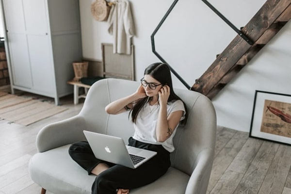 Woman sitting on stylish loveseat working on laptop in trendy loft space with rustic floors and modern design