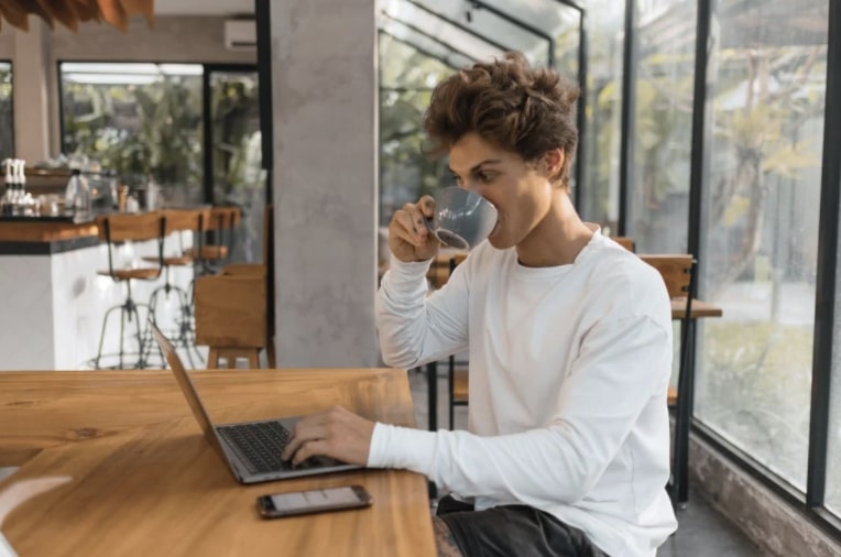 Young man sipping coffee and working on laptop on wooden table in solarium at stylish coworking space