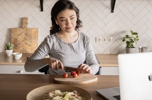 Young woman cutting tomatoes and teaching cooking class in stylish kitchen in front of opened laptop