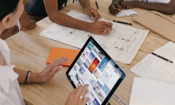 Three people working at table, talking and interacting on tablet while drawing out plans on work table in office