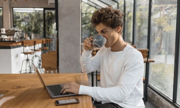 Young man sipping coffee and working on laptop on wooden table in solarium at stylish coworking space