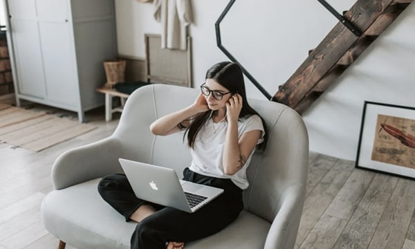 Woman sitting on stylish loveseat working on laptop in trendy loft space with rustic floors and modern design