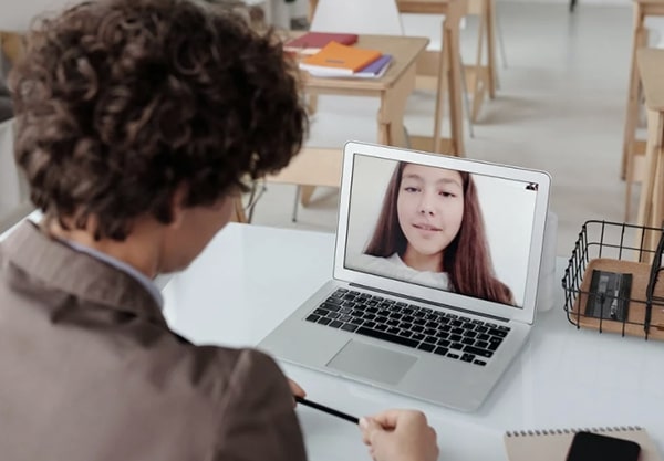 Over the shoulder view of a teacher seated at desk in classroom, chatting with young student on a laptop computer via video conferencing