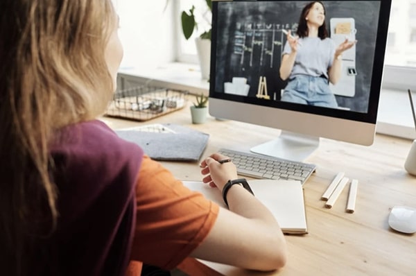 Over the shoulder view of young girl using desktop computer, writing in notebook and watching teacher teaching onscreen