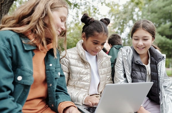 Three young girls sitting outside in park, looking and laughing at a laptop the girl in the middle is holding