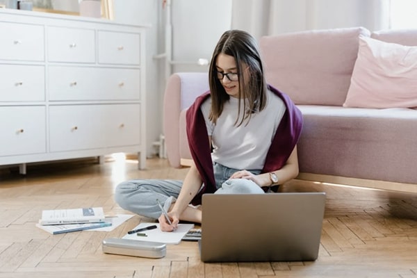 Young woman sitting on floor against couch, at home with open laptop and textbooks, writing in notebook