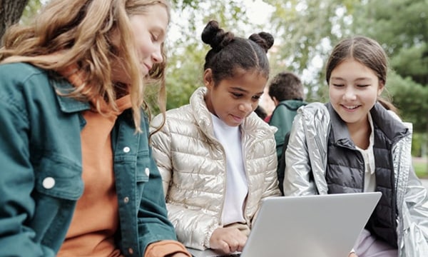 tile Three young girls sitting outside in park, looking and laughing at a laptop the girl in the middle is holding-min