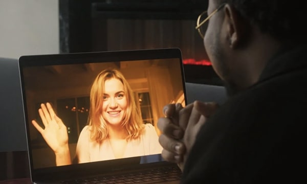 Over-the-shoulder view of man chatting with happy woman waving on-screen of laptop