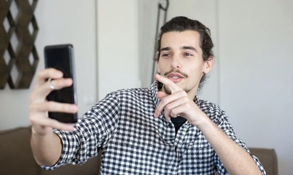 View of man in living room, speaking and interacting with smartphone held at arm’s length whiles gesticulating and pointing finger