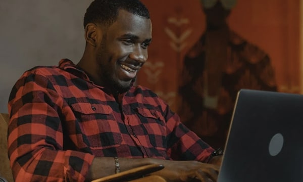 Man seated, facing the right and smiling while engaged and typing on laptop on lap with piece of art in the backgroun