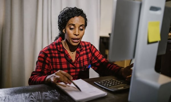 View of woman at home in office in front of desktop looking down and pointing a pen at notebook, while speaking and engaging with screen