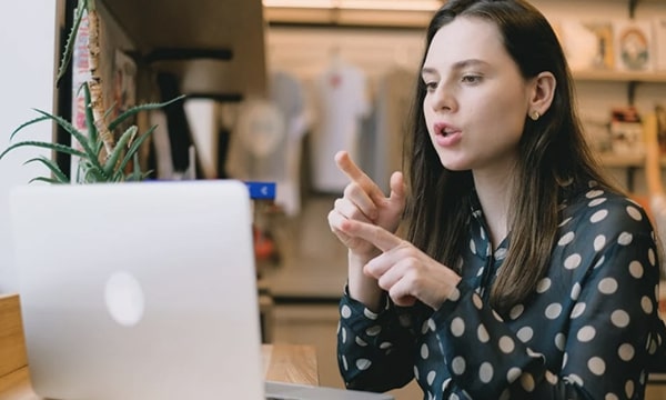 Young woman wearing business casual, moving her hands and speaking in front of laptop on a ledge in a communal workspace