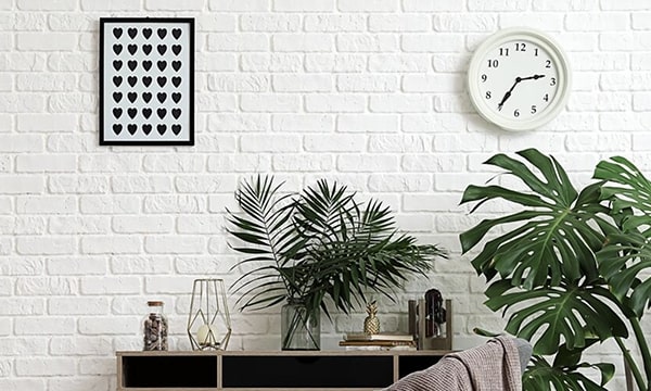 White brick background with art hung on wall, and analog clock, plus plants and a desk with a vase, more plants, books and a paperweight