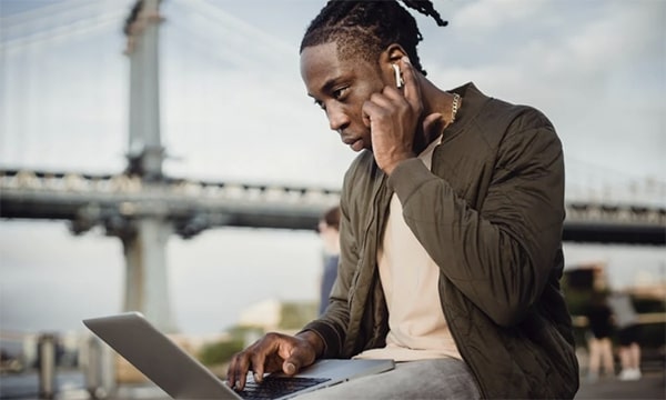 Man outside sitting with a suspension bridge in the background, focused on his laptop while pushing down his ear bud with left hand