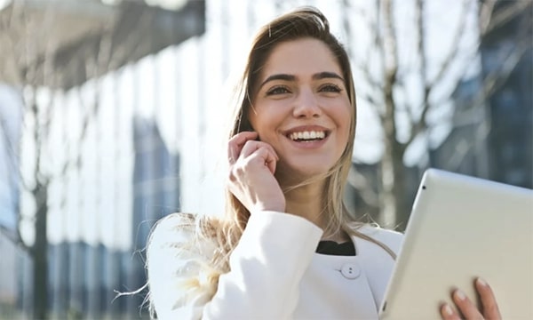 View of smiling business-casual dressed woman outside, holding a phone to her ear and a tablet in the other, while walking and working