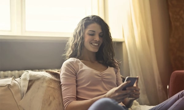 Woman sitting cross-legged, leaning against bed’s headrest, smiling and looking down at her phone in her hands