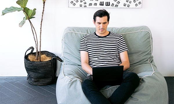 Casual man sitting on bean bag with open laptop in stylish setting with plant to the left and art hanging on the wall
