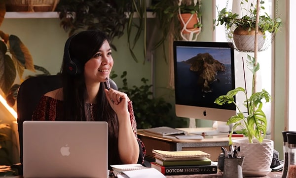 Smilende, kontemplativ kvinde, der arbejder hjemmefra med bærbar computer og desktop, iført hovedtelefoner ved skrivebordet, omgivet af planter