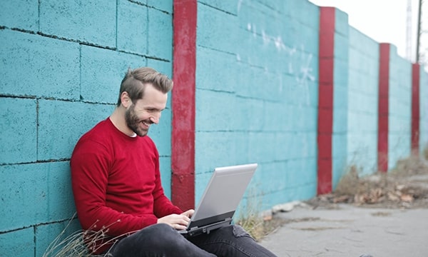 Smiling man sitting outside, leaning against teal brick wall with laptop open on lap, typing and interacting with screen